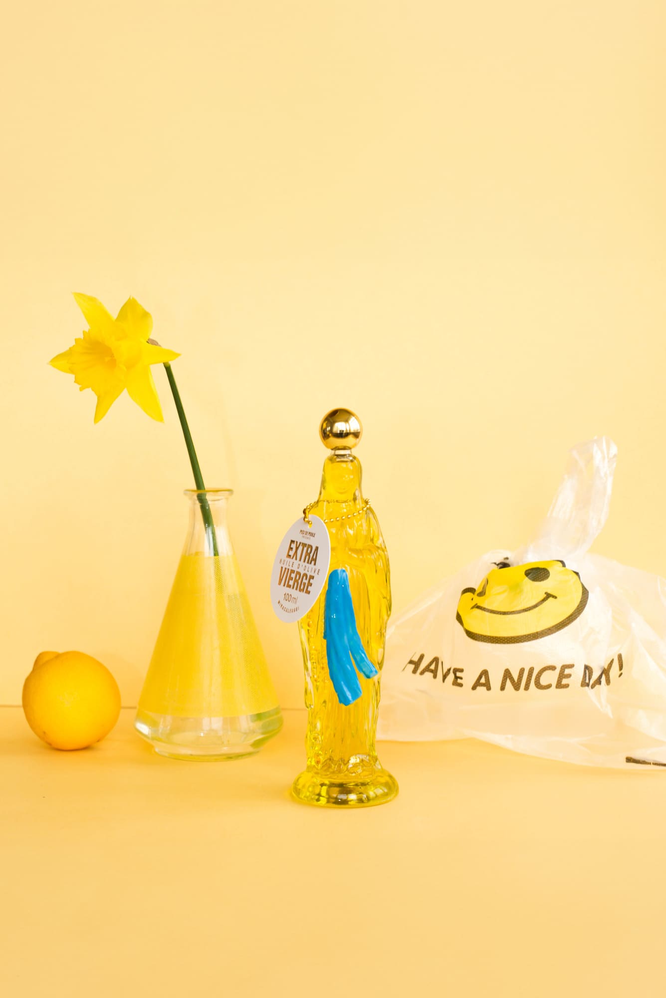 Yellow bottle with a smiley face label, lemon, vase with yellow flower, and 'Have a Nice Day' bag on a yellow background