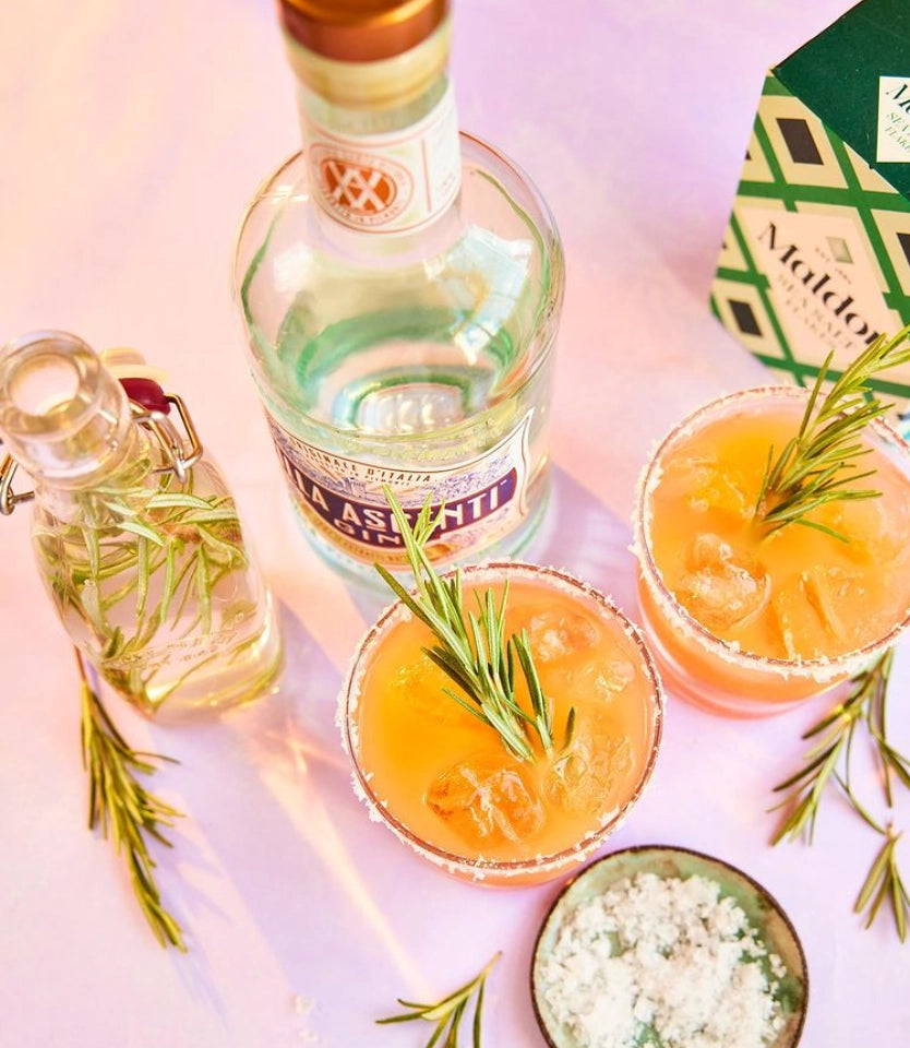 Two cocktails with rosemary garnish and a bottle of Absinthe on a light pink background.