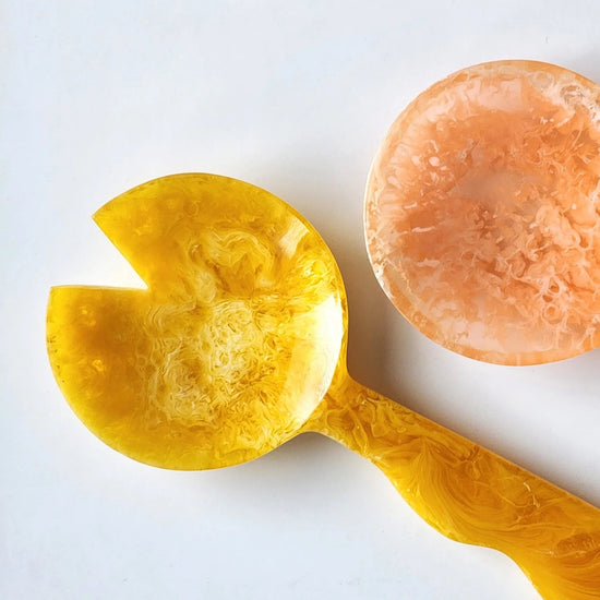 Two decorative spoons, one yellow and one peach-colored, on a white background.