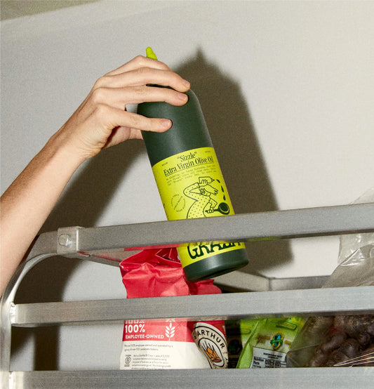 Hand reaching into a refrigerator to grab a bottle of olive oil.