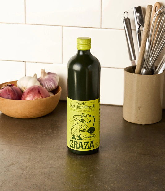 Bottle of Graza olive oil on a kitchen counter with ingredients and utensils in the background.