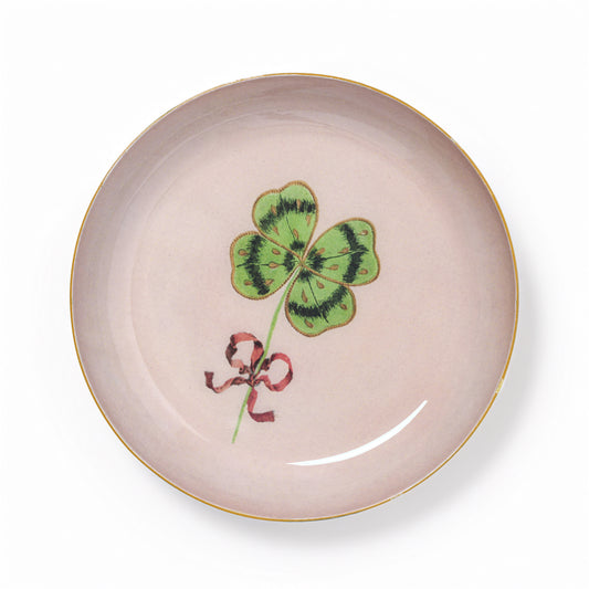 A round light pink ceramic plate with a gold rim, featuring a green four-leaf clover and a red ribbon illustration in the center, set against a plain white background.