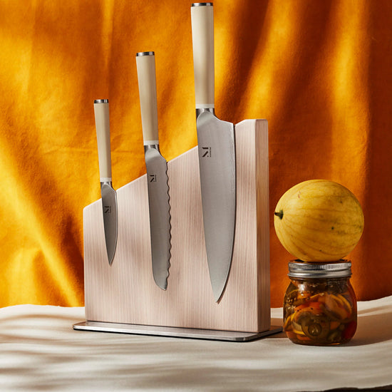 Set of knives on a wooden magnetic knife stand with a yellow fruit and jar on a marble surface.