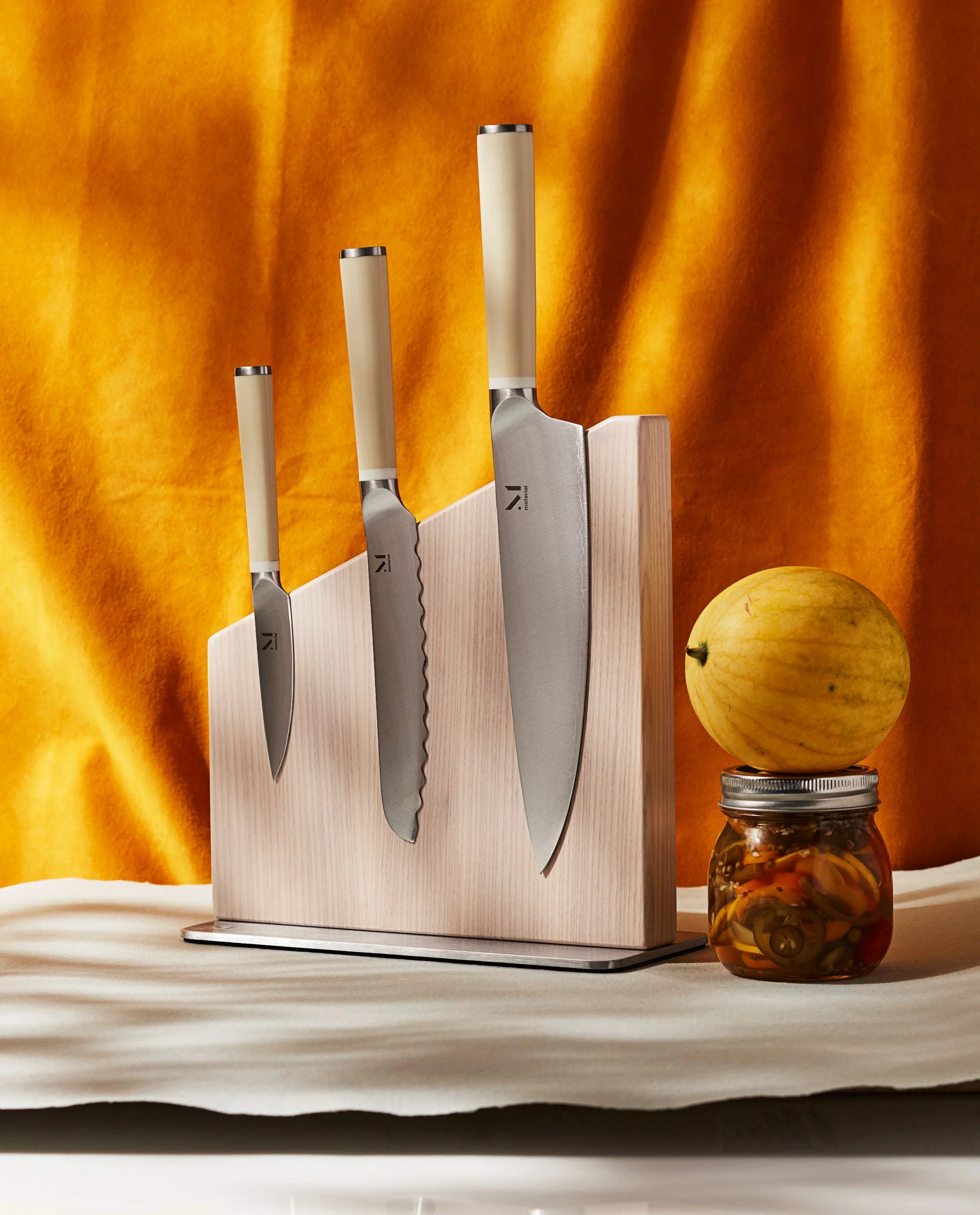Set of knives on a wooden magnetic knife stand with a yellow fruit and jar on a marble surface.