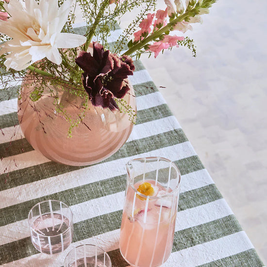 Pink floral arrangement in a vase on a striped tablecloth with glasses and a Clear water carafe