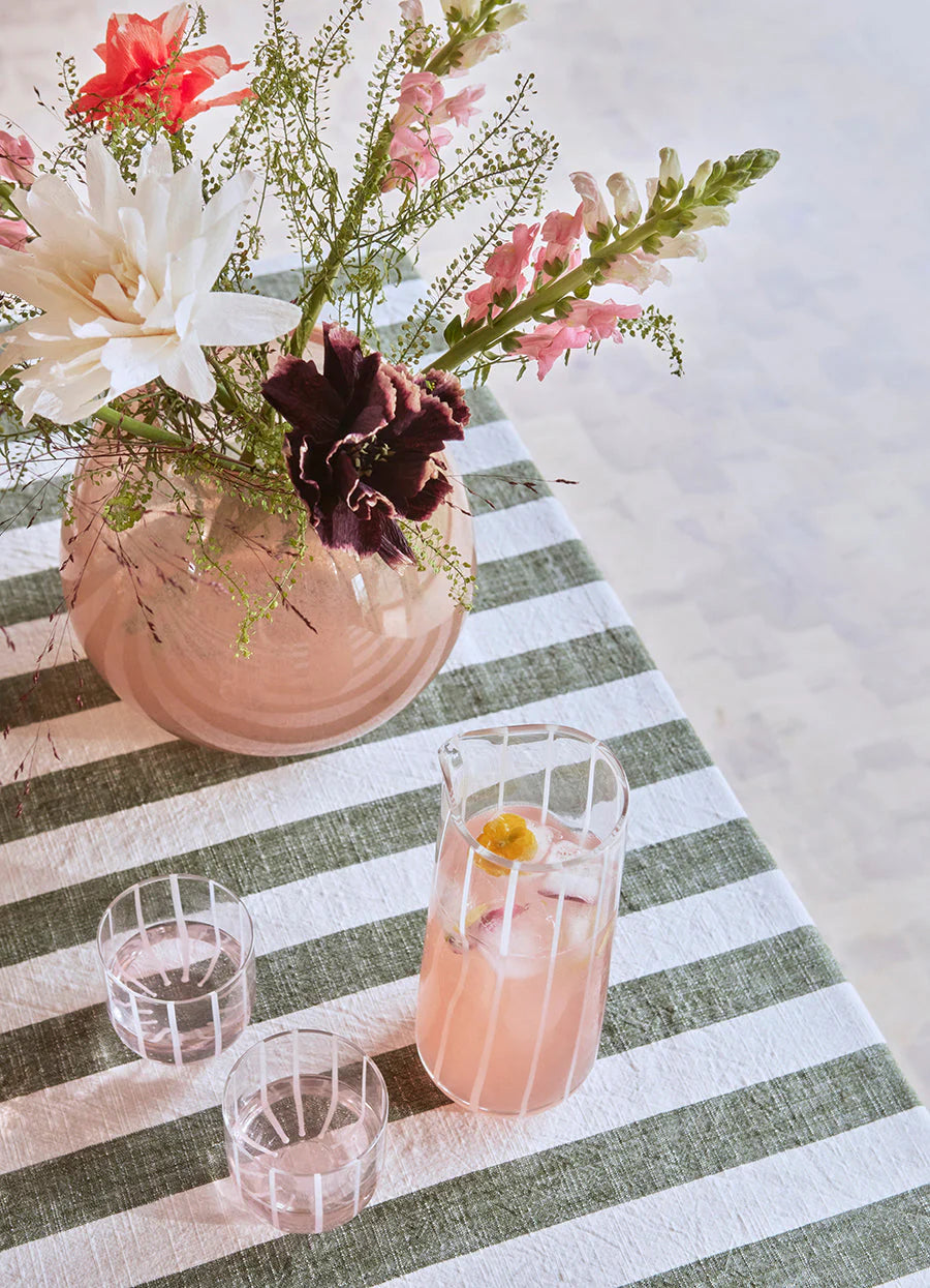 Pink floral arrangement in a vase on a striped tablecloth with glasses and a Clear water carafe