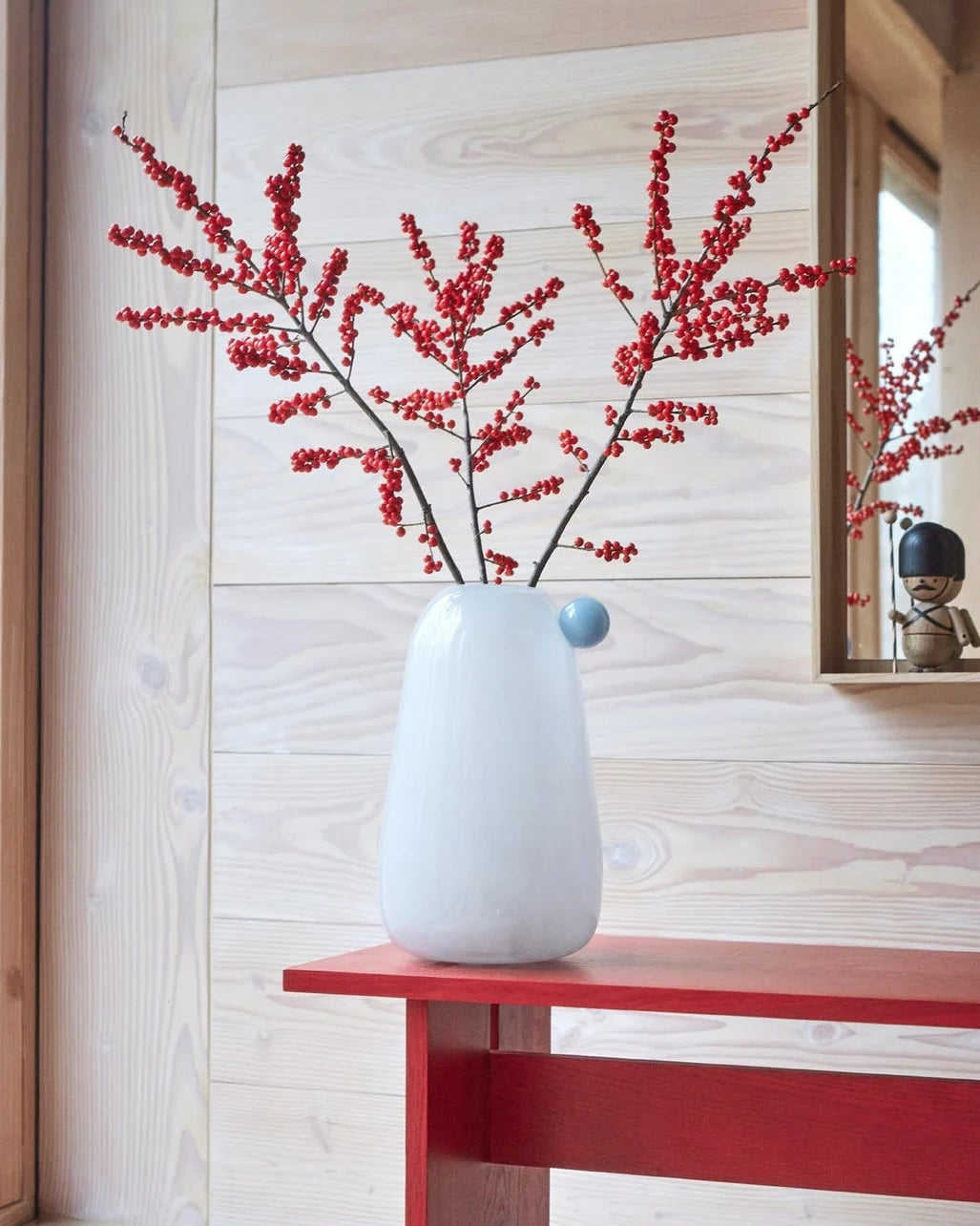 White vase with red branches on a red bench against a wooden wall.