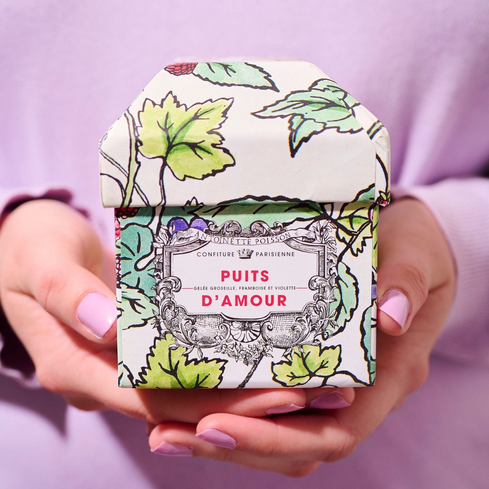 A person holding a small decorative box with a floral pattern and the label “Puits d’Amour” in pink text