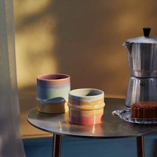 Two ceramic pots with gradient colors on a metal table next to a coffee maker and cake.