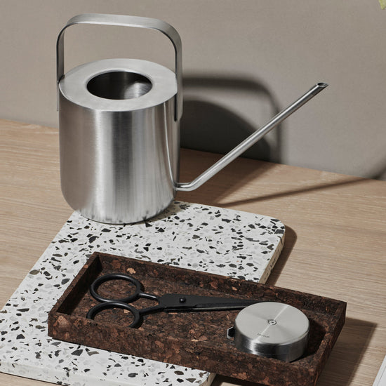 Silver watering can on a wooden table with a tray, scissors, and a candle.