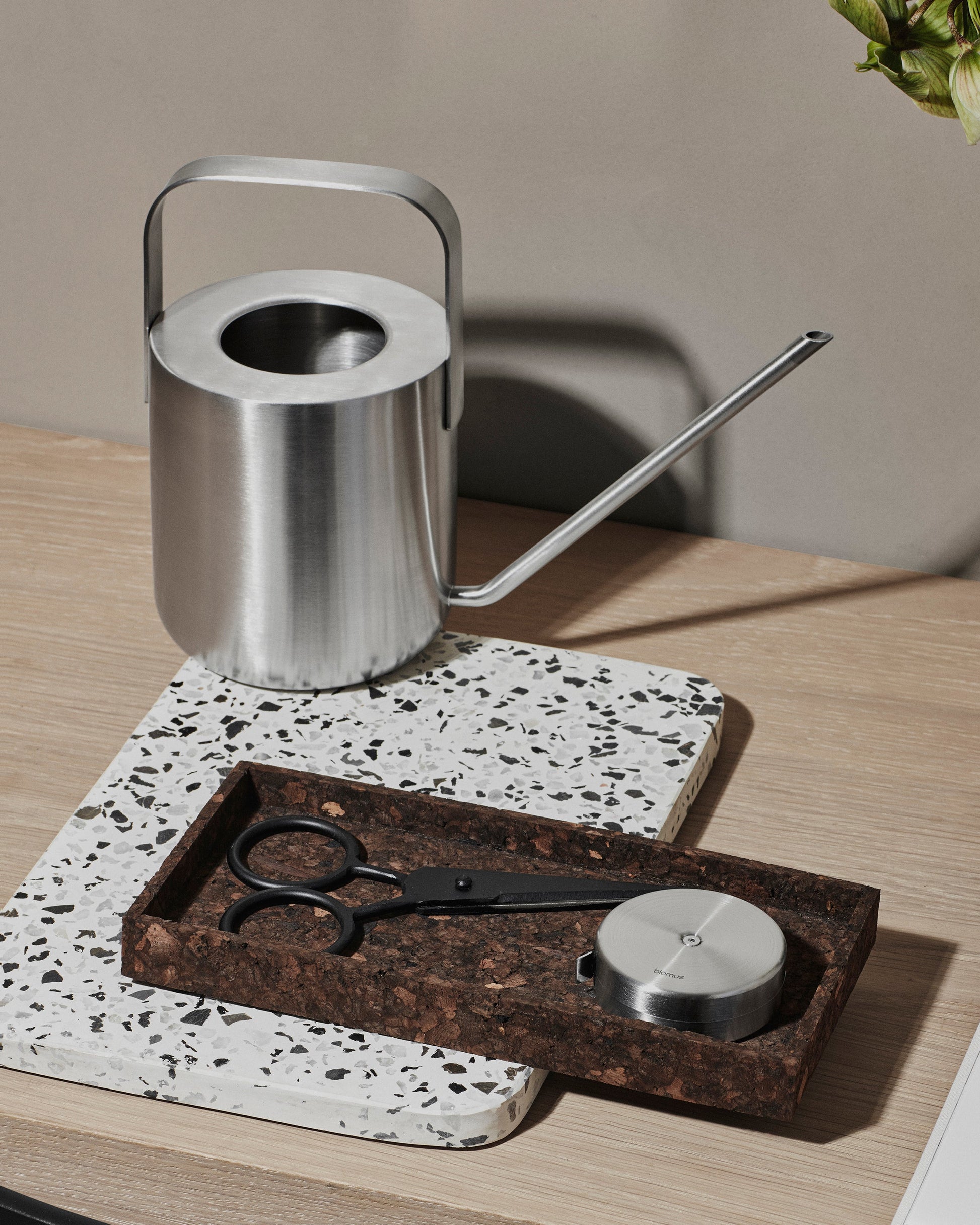 Silver watering can on a wooden table with a tray, scissors, and a candle.
