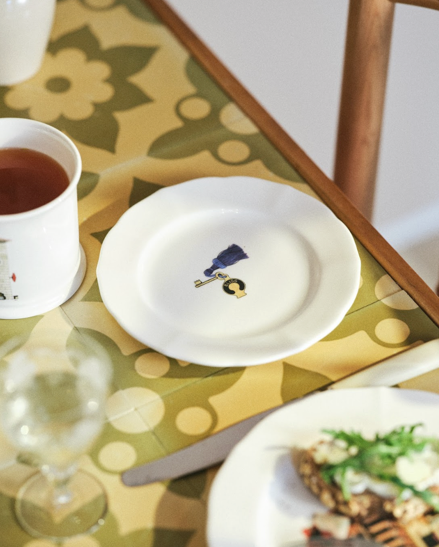 White plate with a design on a patterned tablecloth, next to a cup of tea and a glass.