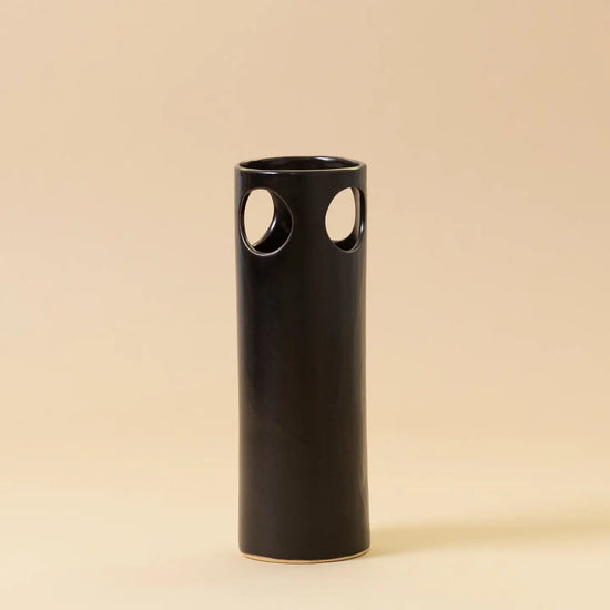 Black cylindrical vase with circular cutouts on a beige background