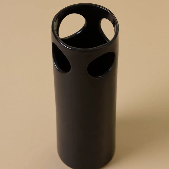 Black cylindrical vase with circular holes on a beige background