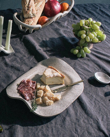 Table setting with cheese platter, bread, fruits, and cutlery on a dark surface.