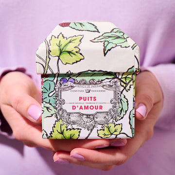 A person holding a small decorative box with a floral pattern and the label “Puits d’Amour” in pink text