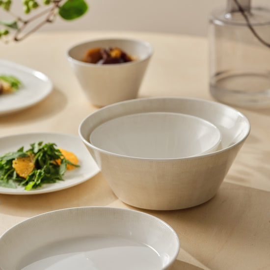 White ceramic bowls on a light wooden surface with blurred plates of food in the background.