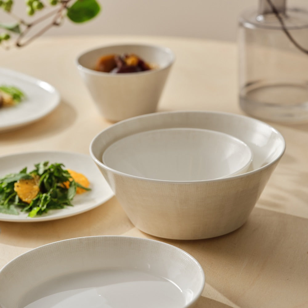 White ceramic bowls on a light wooden surface with blurred plates of food in the background.