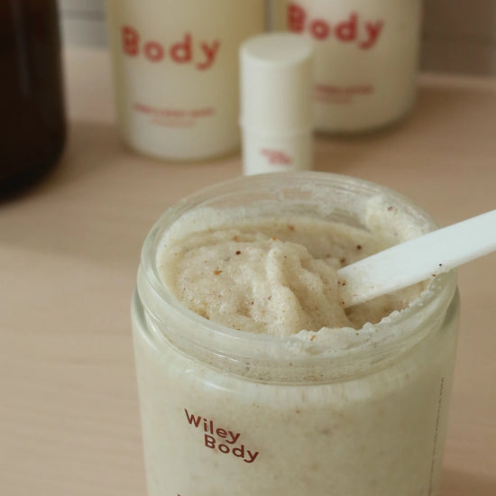 A jar of Wiley Body Body Scrub filled with a light beige exfoliating scrub, shown on a wooden surface with other Wiley Body products in the background.