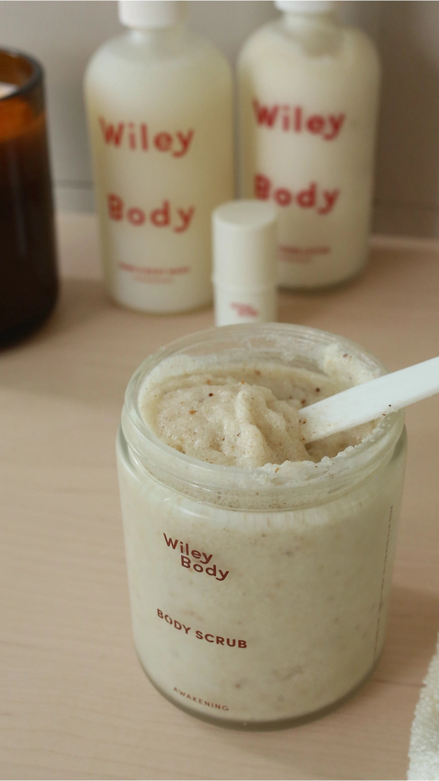 A jar of Wiley Body Body Scrub filled with a light beige exfoliating scrub, shown on a wooden surface with other Wiley Body products in the background.