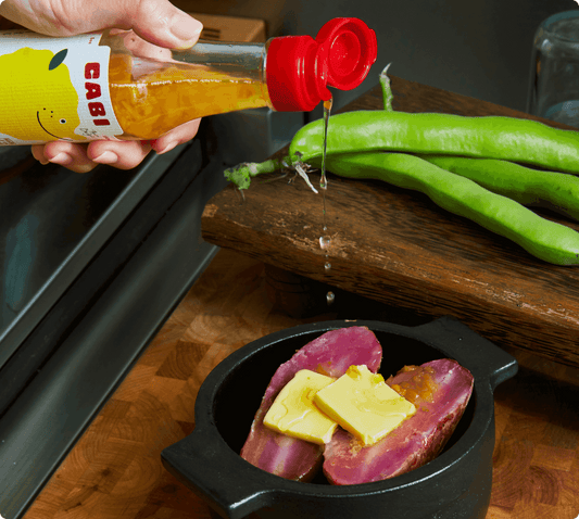 Person pouring a sauce from a 'Cabi' bottle onto food in a pot on a wooden surface.