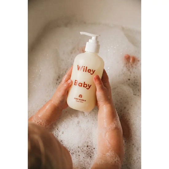 A person holding a frosted bottle labeled 'Viley Baby' with red text, surrounded by foamy bath water in a bathtub.