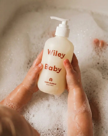 A person holding a frosted bottle labeled 'Viley Baby' with red text, surrounded by foamy bath water in a bathtub.