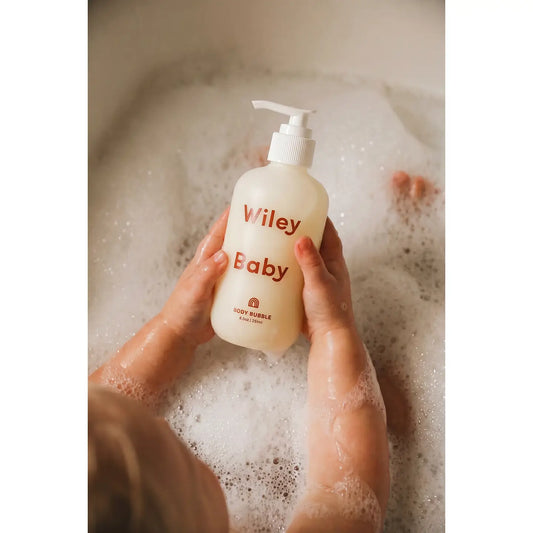 A person holding a frosted bottle labeled 'Viley Baby' with red text, surrounded by foamy bath water in a bathtub.